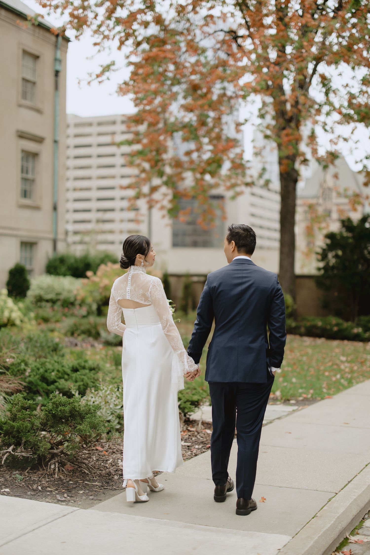 Bride in a white dress and groom in a navy suit hold hands while walking down a sidewalk lined with autumn trees and gardens.