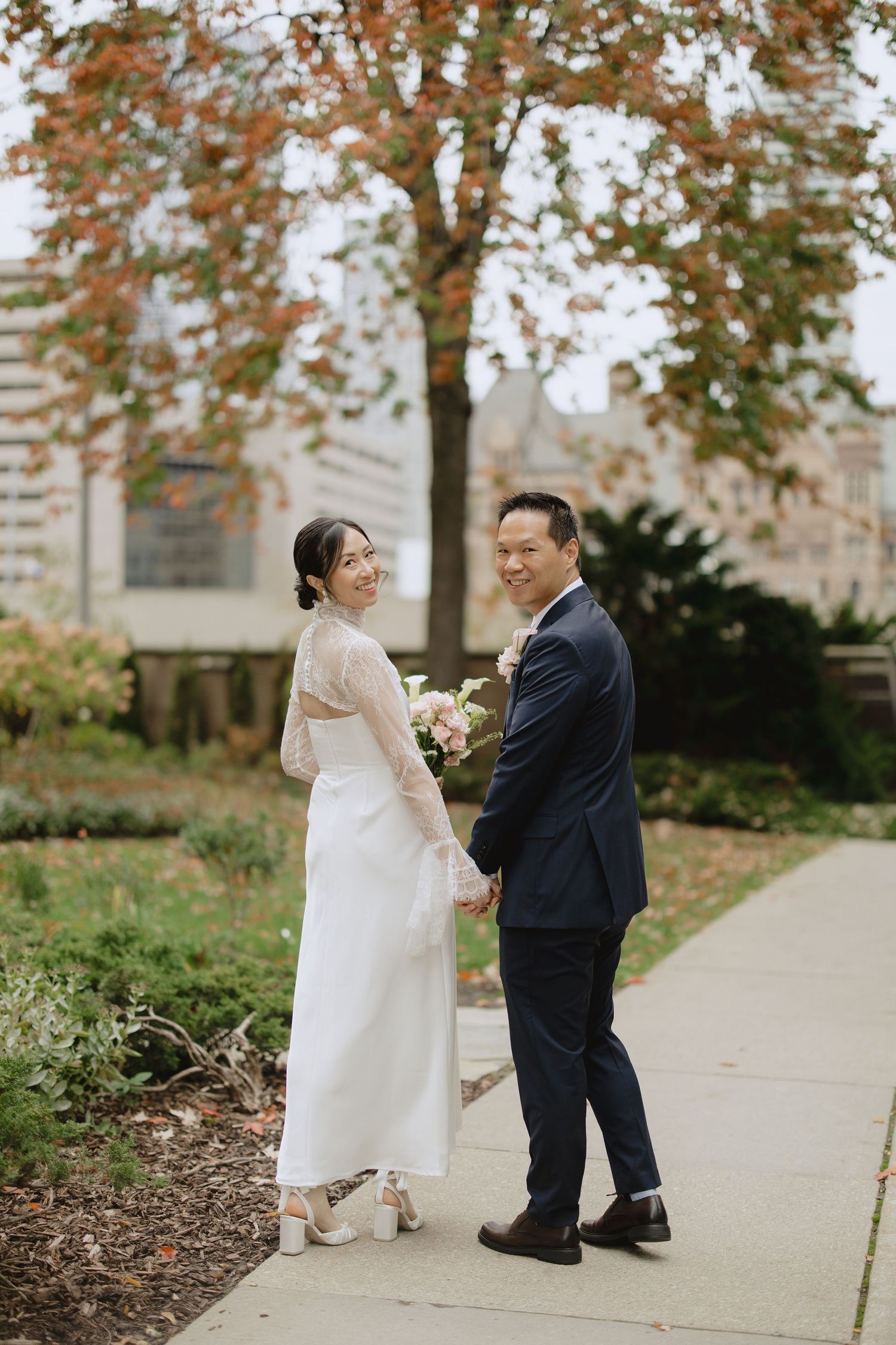 Bride in a white long-sleeve gown holding a bouquet and groom in a navy suit smile back at the camera on a path beneath an autumn tree.