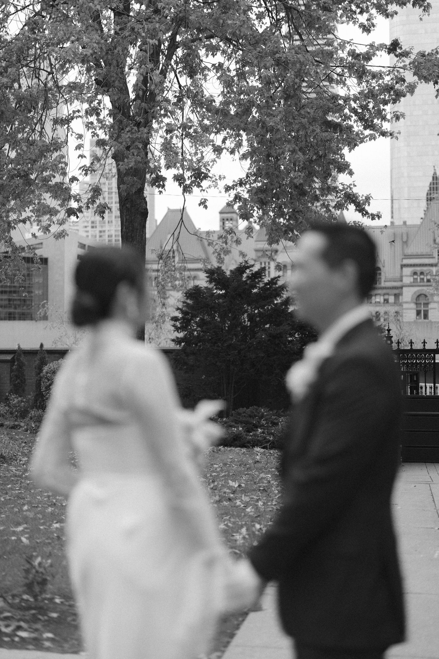Black and white photo of a blurred bride and groom holding hands in a park, with historic buildings and autumn trees in the background.