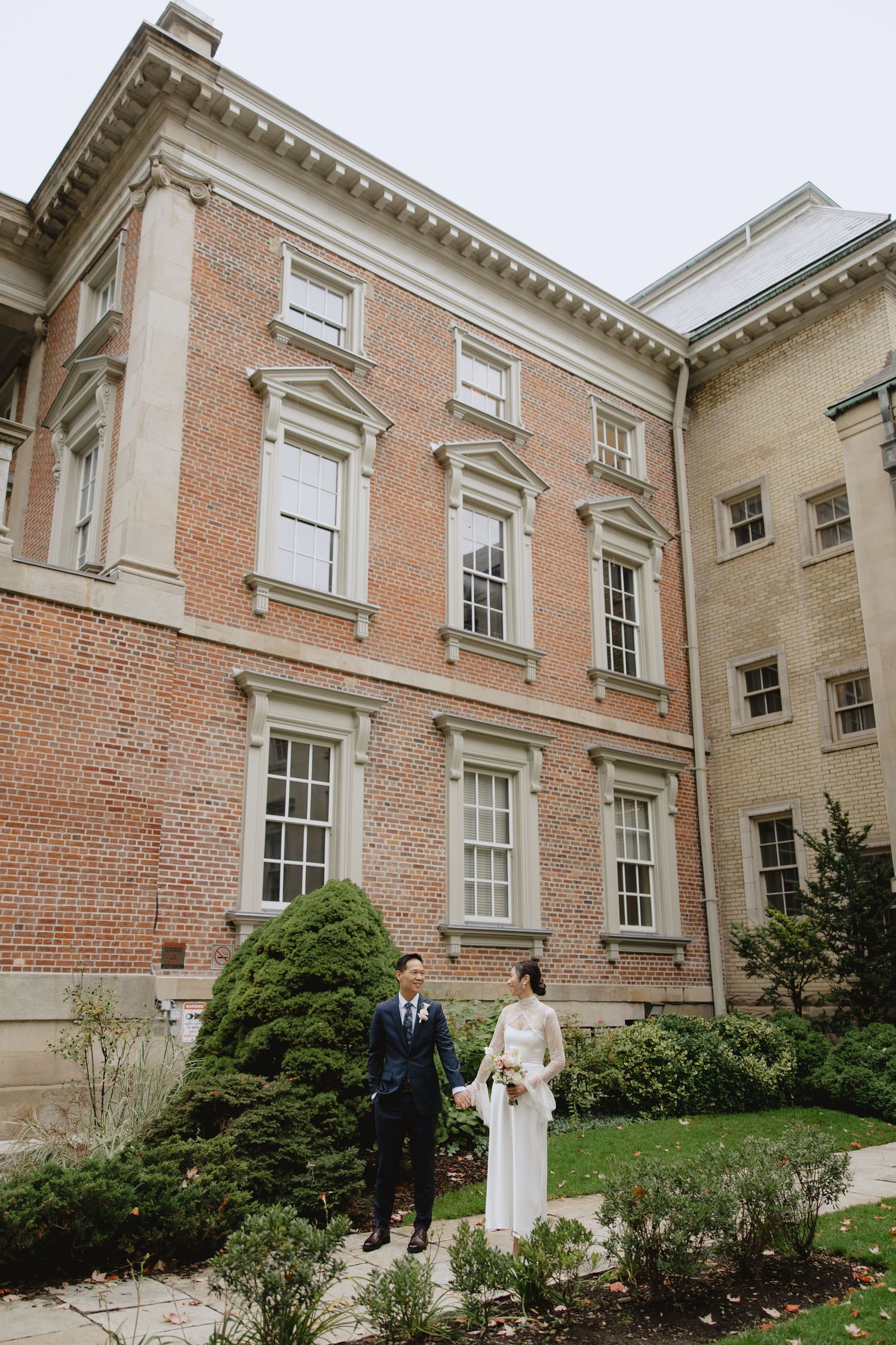 A bride and groom stand hand in hand on a stone path beside a red brick building framed by evergreen shrubs.