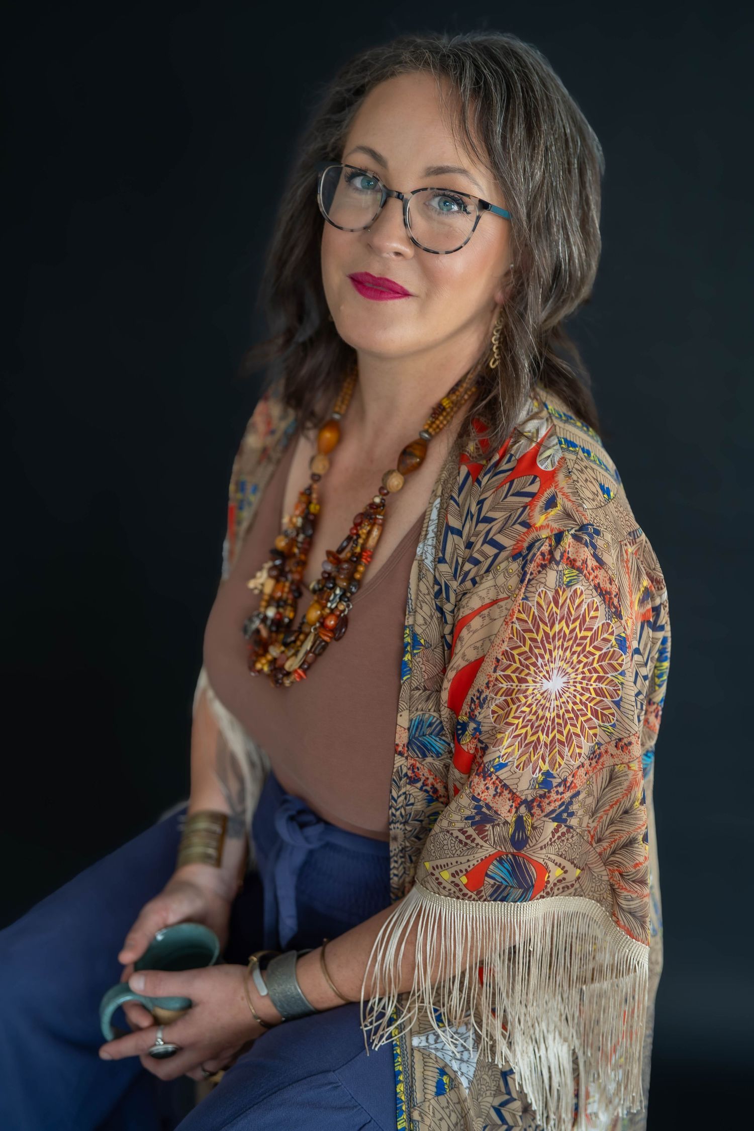 Woman with glasses and red lipstick wearing a colorful fringed kimono, beaded necklace and bangles, seated against a dark backdrop.