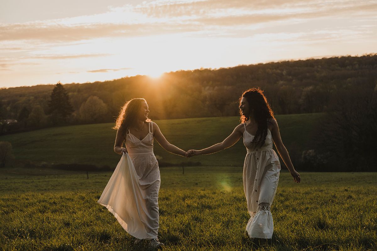 Two women in light flowing dresses hold hands in a grassy field at sunset, with rolling hills and glowing sky behind them.