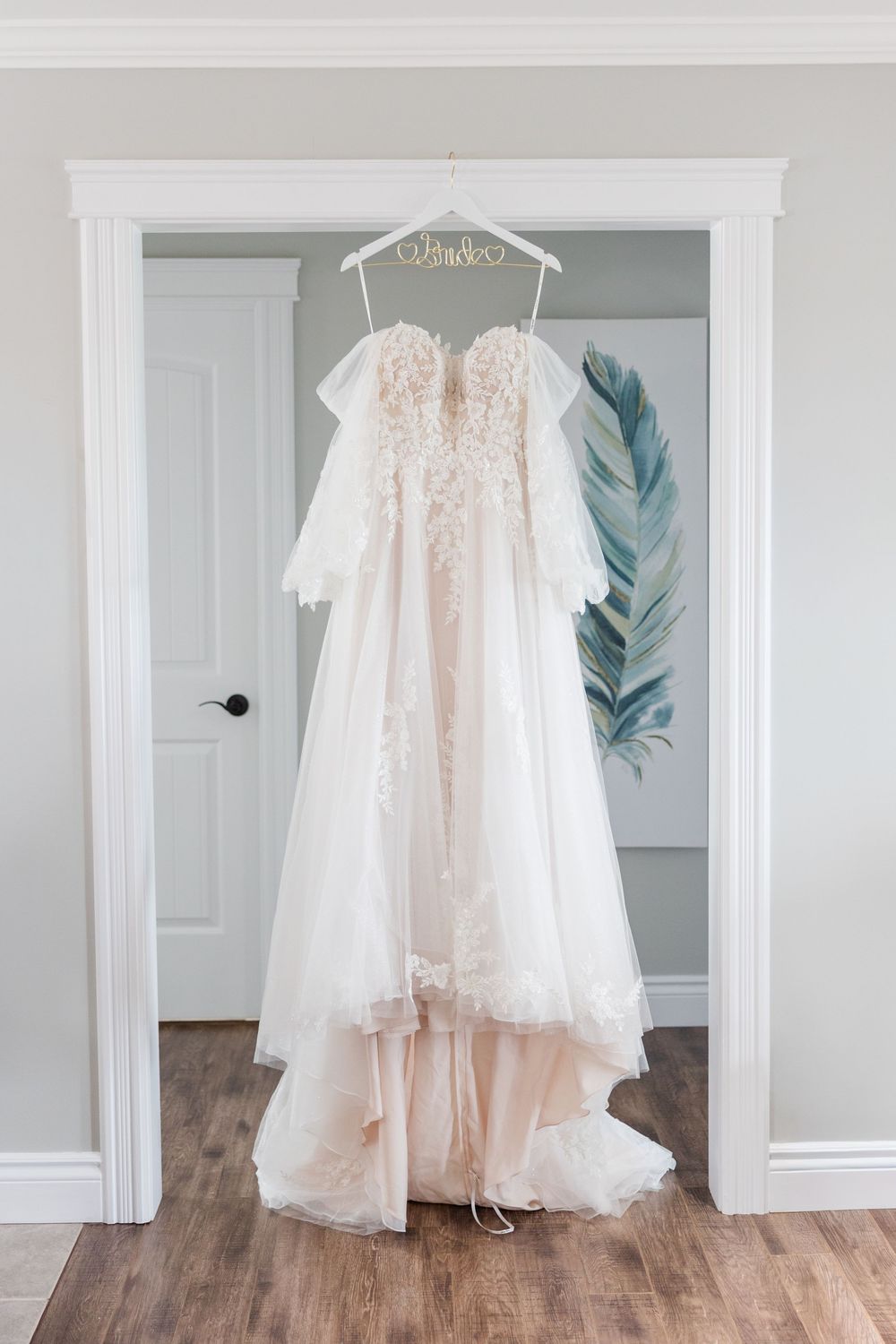 Elegant blush lace wedding dress hanging on a personalized bridal hanger in a bright white doorway.