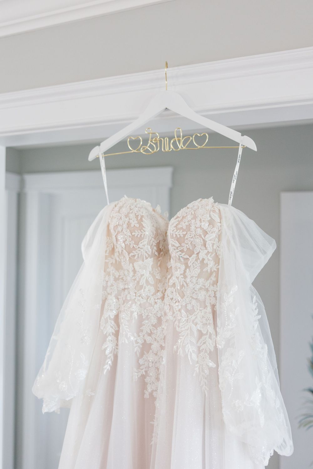 A blush lace strapless wedding gown hanging on a white bride hanger with gold lettering in a bright elegant room.