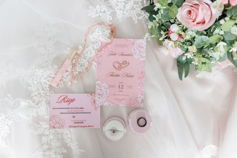 Pink floral wedding invitation suite with RSVP card, diamond rings, lace details, and fresh roses on white fabric backdrop.