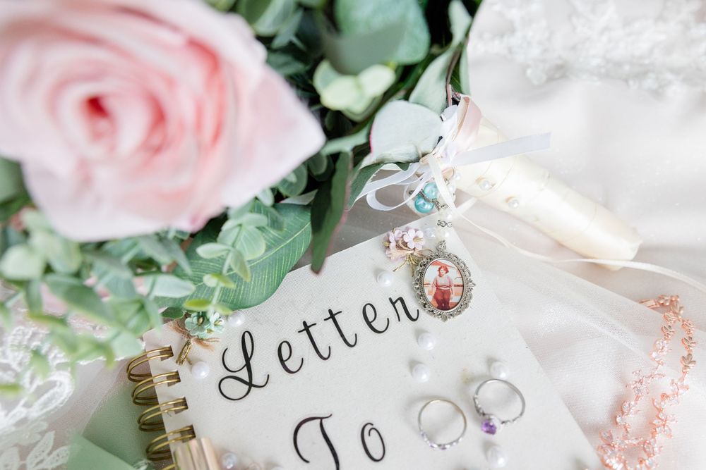Wedding flat lay featuring a pink rose, eucalyptus, rings, charm bracelet, and a letter book on white fabric.