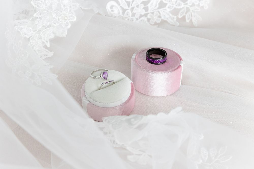 Two wedding rings in pink velvet boxes on white lace bridal veil, featuring diamond engagement ring and purple gemstone band.