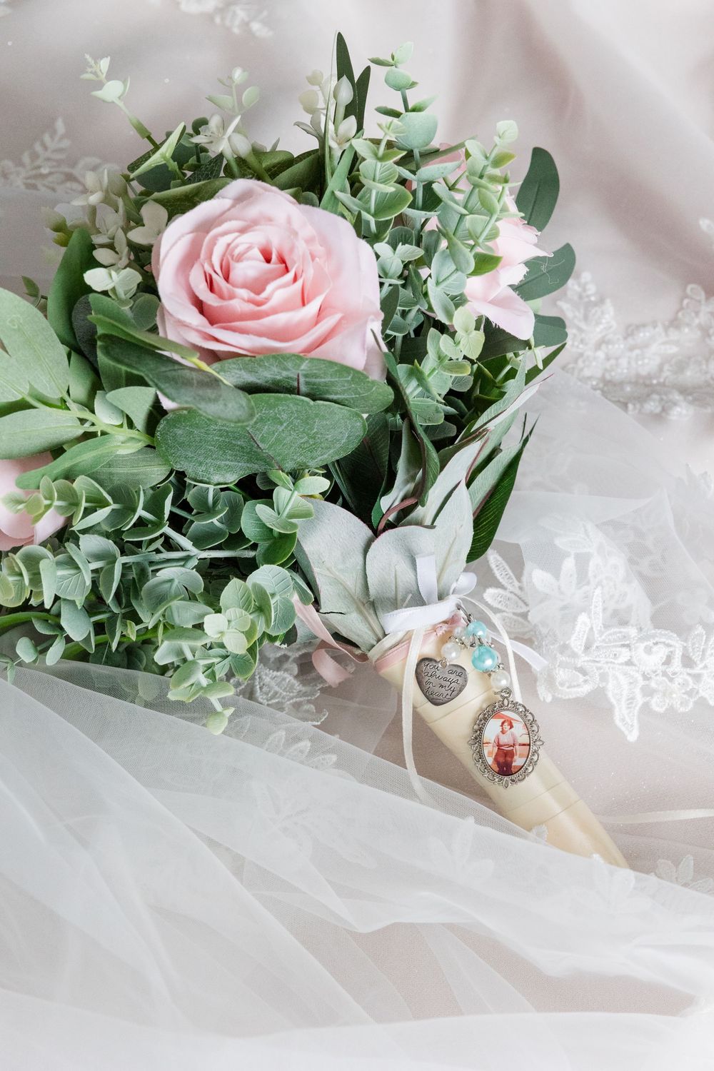 Bridal bouquet with pink roses and eucalyptus greenery, adorned with a charm and ribbon, resting on white lace fabric.