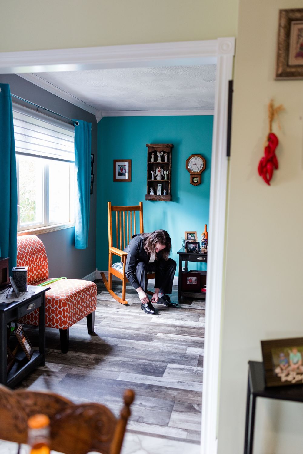 Person tying shoes in a cozy living room with teal walls, hardwood floors, and a wooden rocking chair near a bright window.