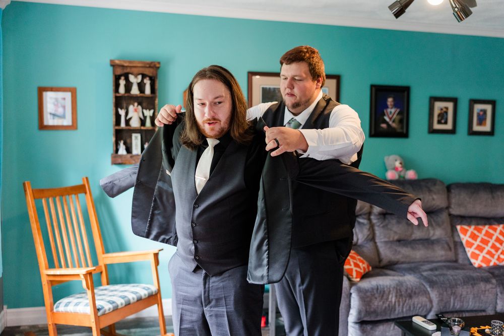 Two men in formal suits help each other get dressed in a living room before a special event.