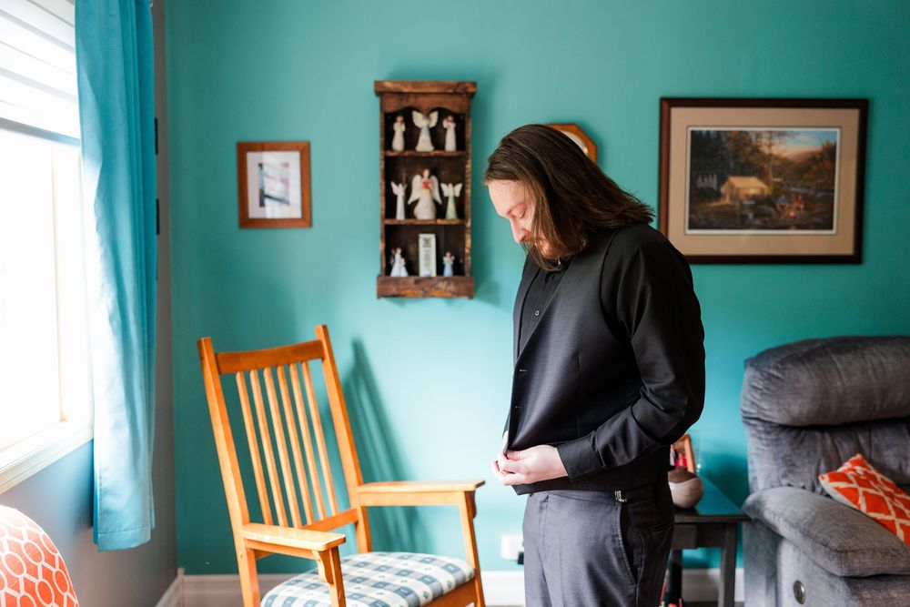 A person in black clothing adjusts their outfit while standing in a teal-walled room with wooden chair and framed art.