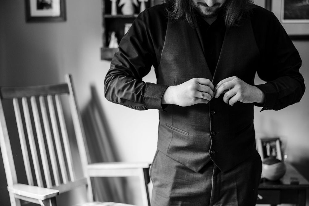 Black and white photo of a groom buttoning his dark dress shirt while getting ready for his wedding day.
