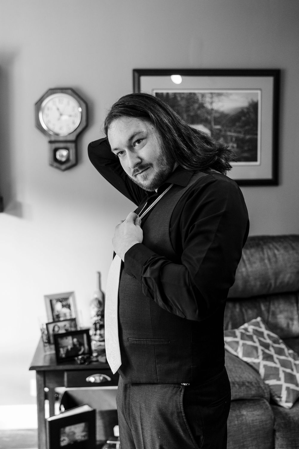 Black and white portrait of a long-haired man adjusting his tie in a suit, standing in a cozy living room.