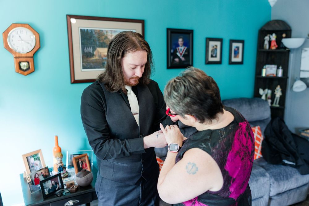 Man with long hair in black jacket helping woman with tattoo in teal living room.