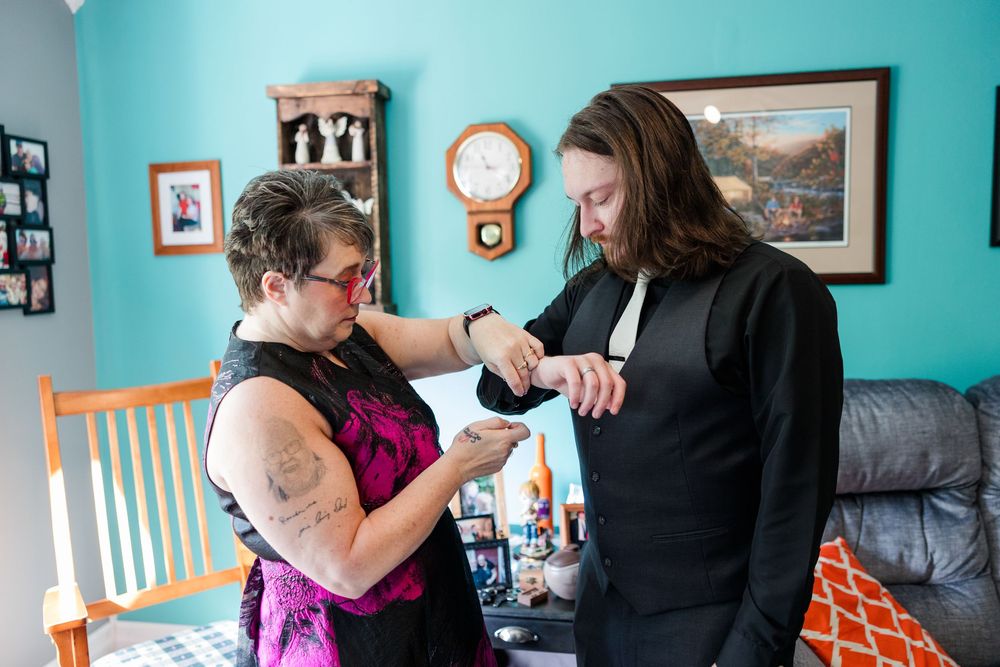 Two women helping each other get dressed for a formal event in a teal living room with a clock on the wall.