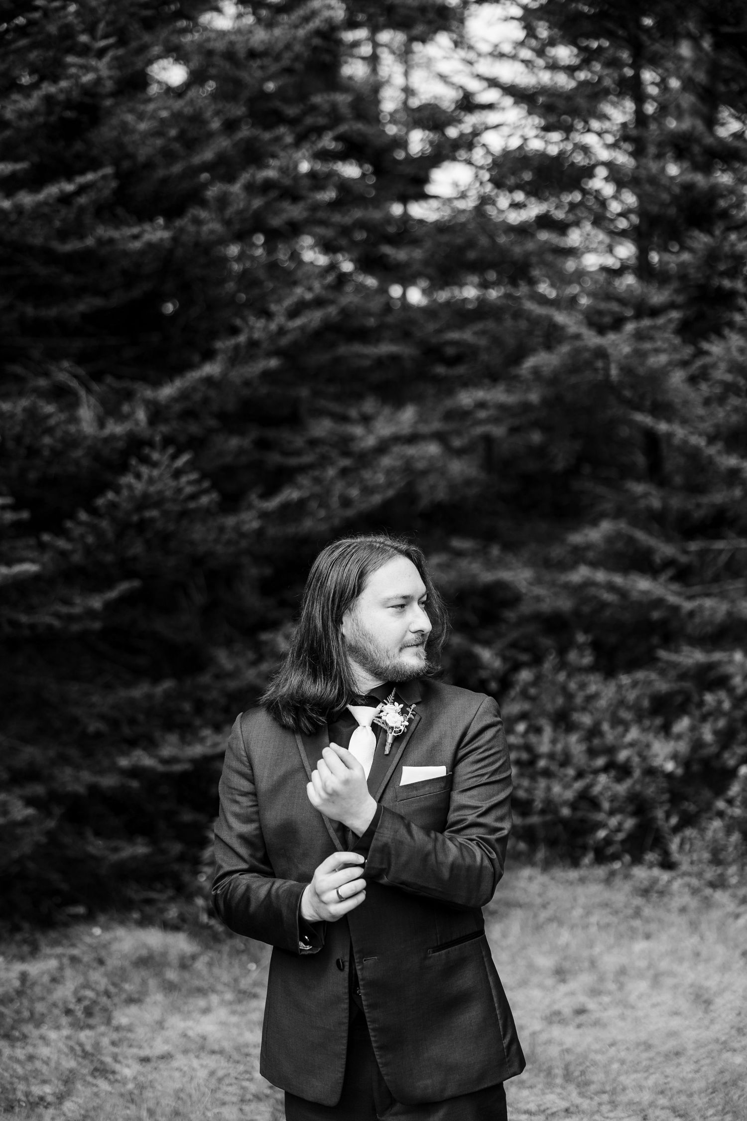 Black and white portrait of a groom in a suit adjusting his boutonniere outdoors among evergreen trees.