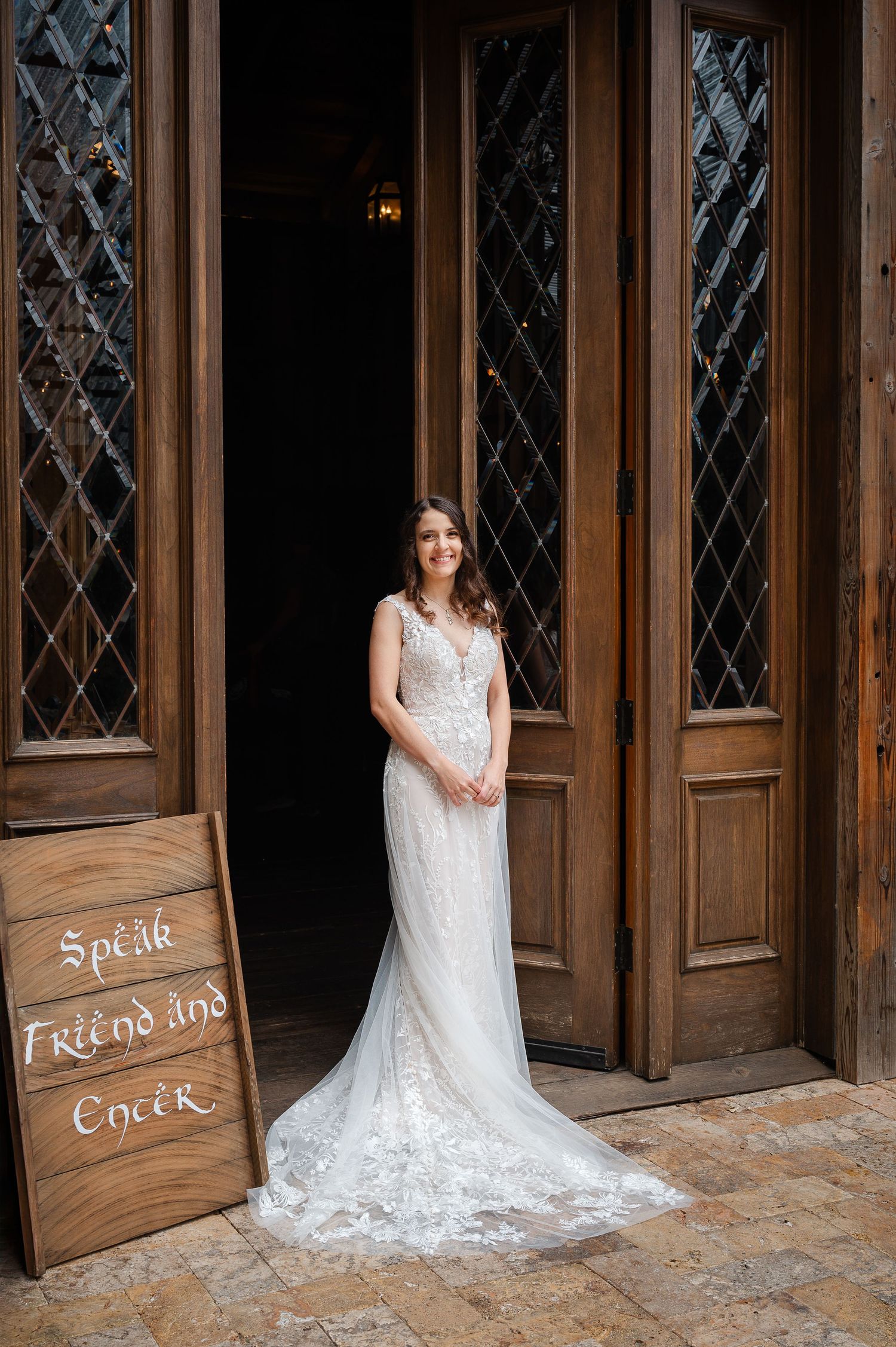 Bride in lace wedding gown stands on wooden deck with waterfall and redwood trees in background.
