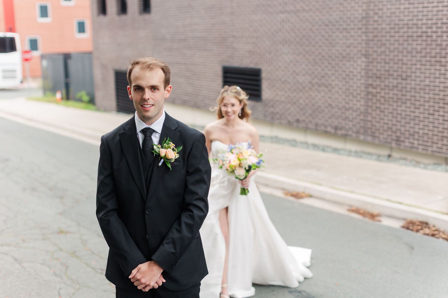 Groom in black tuxedo stands in foreground as bride in white strapless gown holding bouquet approaches on urban street.