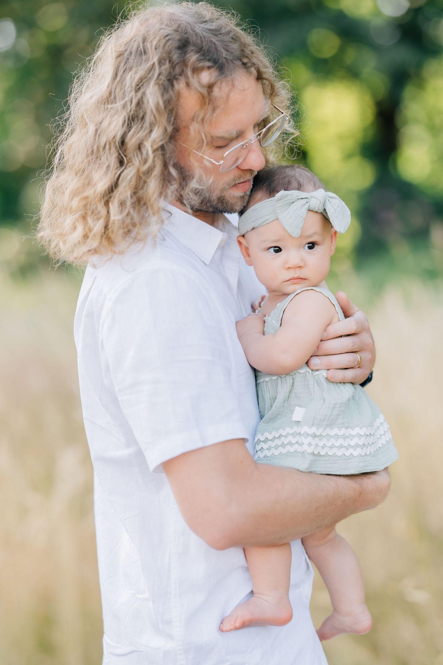 Father with long curly hair holding his baby girl outdoors in a sunny field.