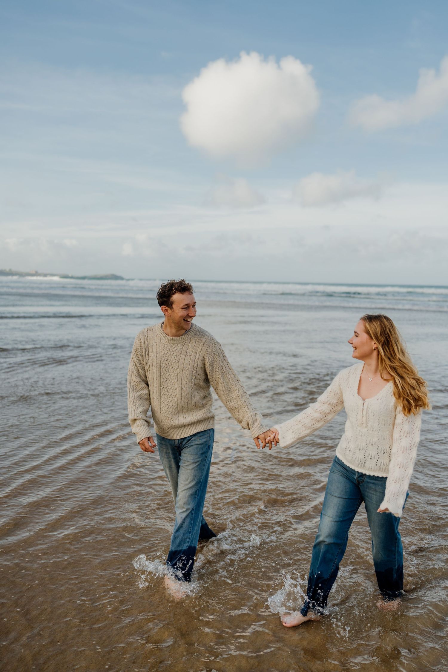 Couple holding hands and walking through ocean waves at watergate bay, wearing cozy knit sweaters and jeans under a cloudy sky.