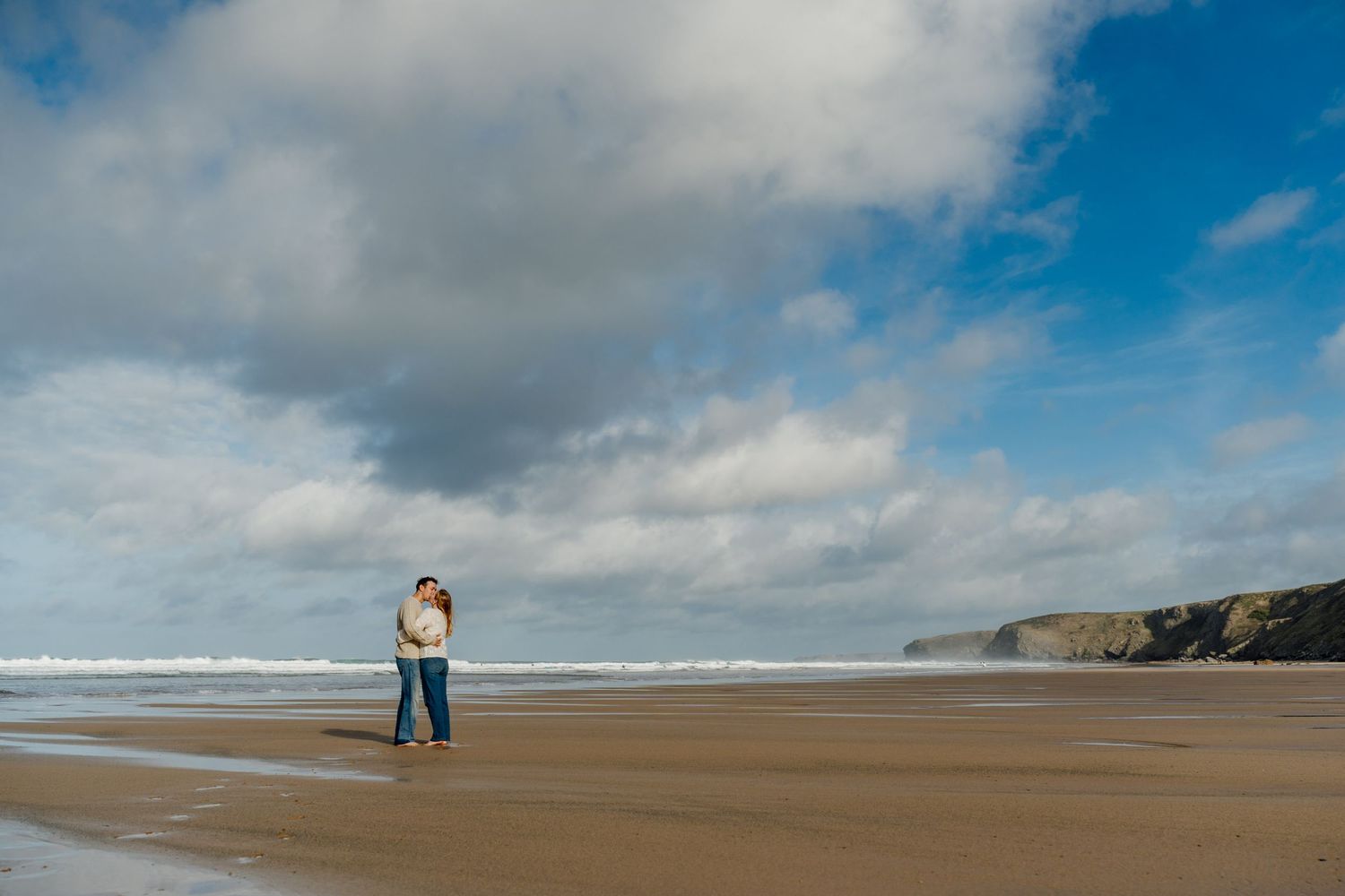 Couple embracing at watergate bay with dramatic cloudy sky and coastal cliffs in the background.