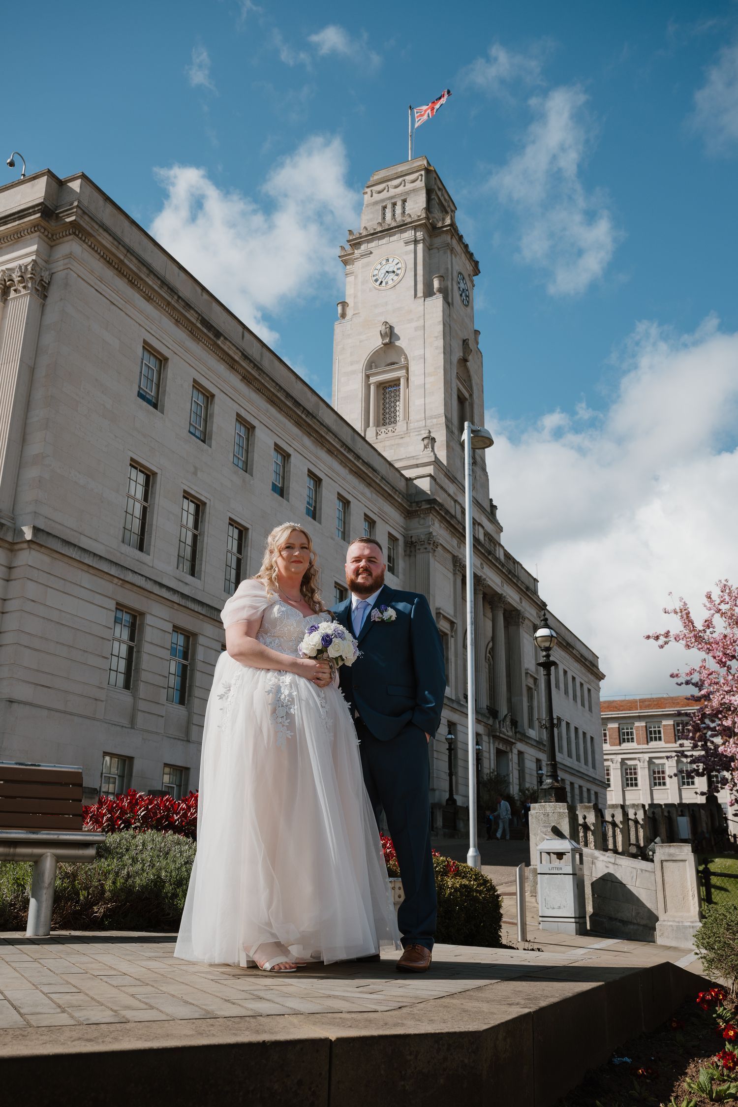 Bride and groom posing outside a grand clock tower building on their wedding day under a blue sky.
