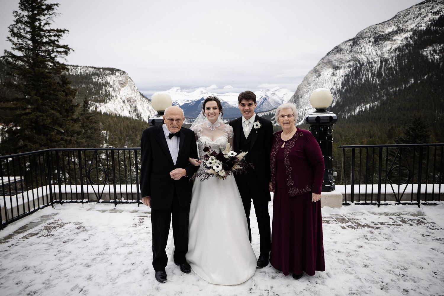 J + R with two loved ones on a snowy Fairmont Banff Springs terrace with snow-capped mountains behind them