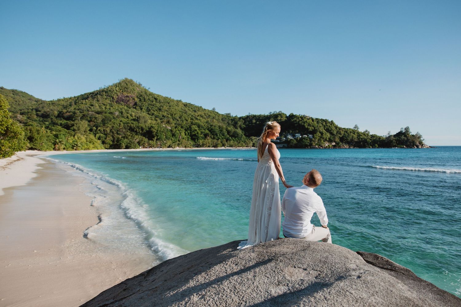 Couple in wedding attire standing on rocks by a turquoise beach with lush green hills in the background.