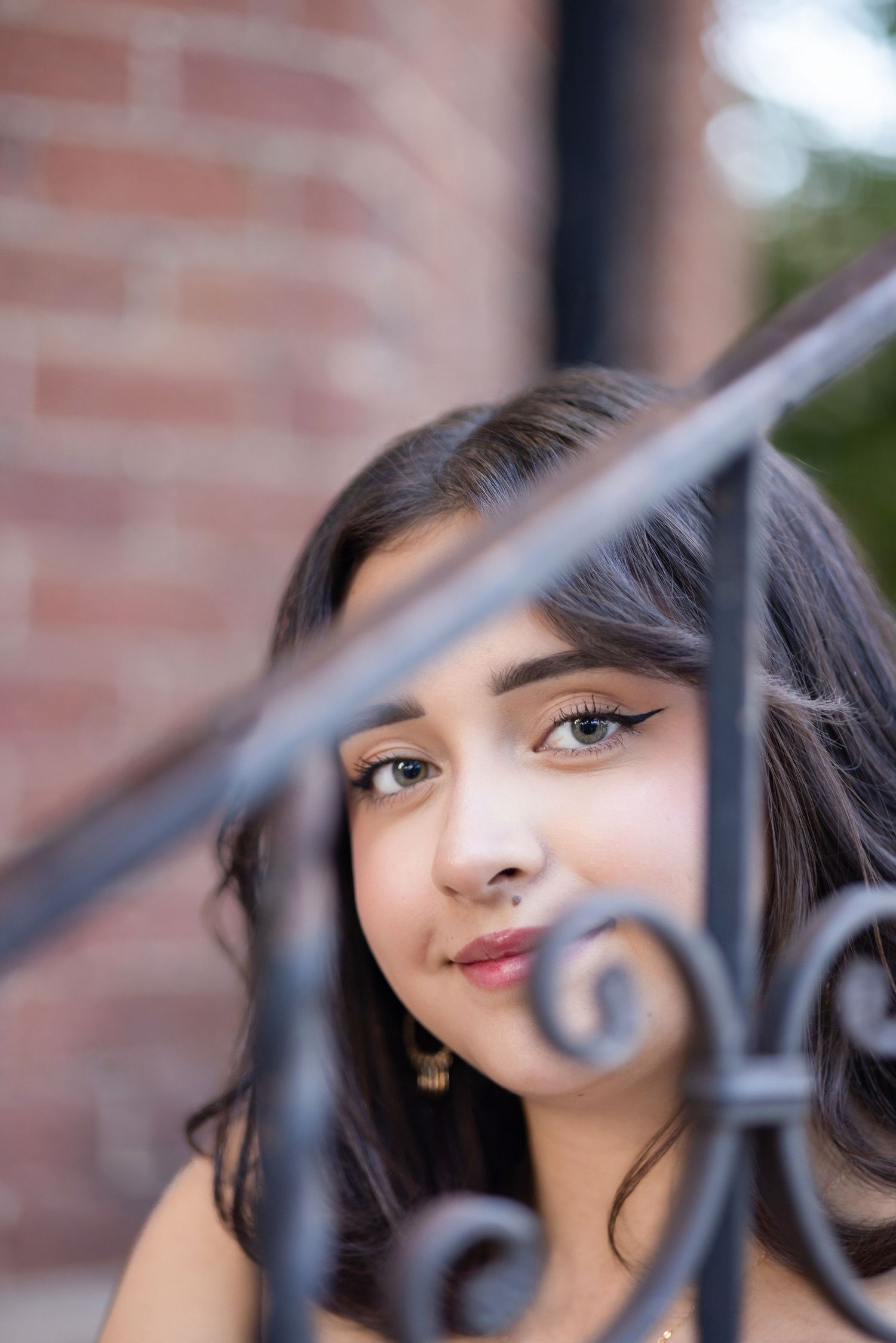 Young woman with dark hair and nose ring gazes through ornate black iron railing, brick wall background.