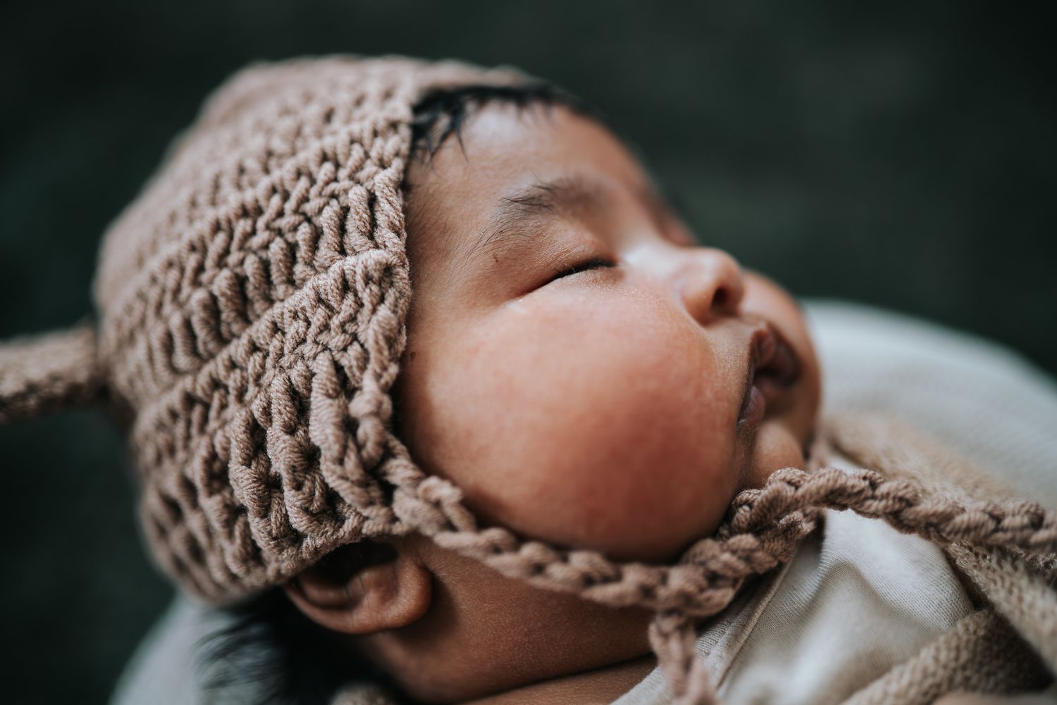 Sleeping newborn baby wearing a cozy knitted brown bonnet hat with tie strings with chubby cheeks in Melbourne.