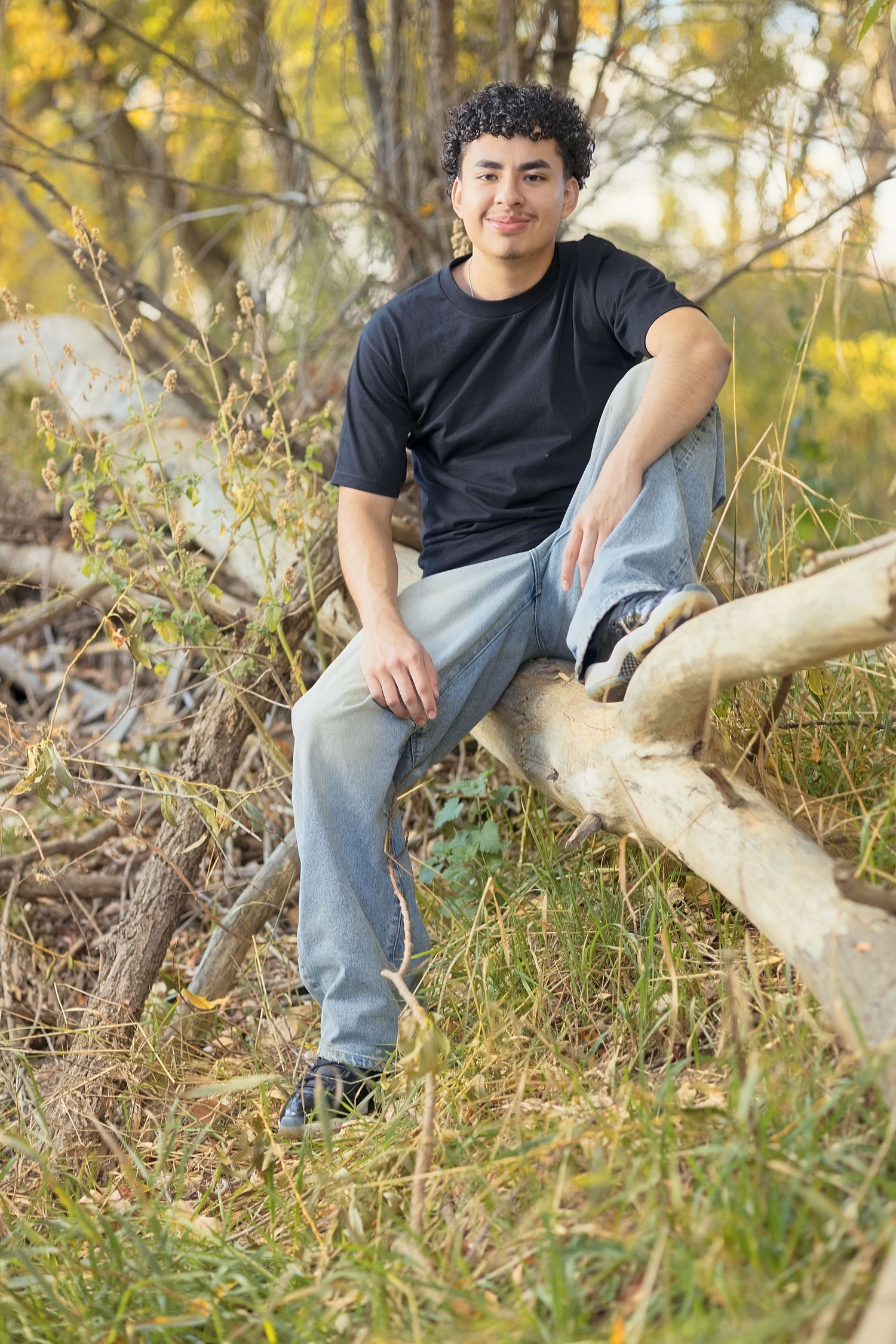 Young man in black t-shirt and jeans sitting on fallen tree branches in a natural outdoor setting.