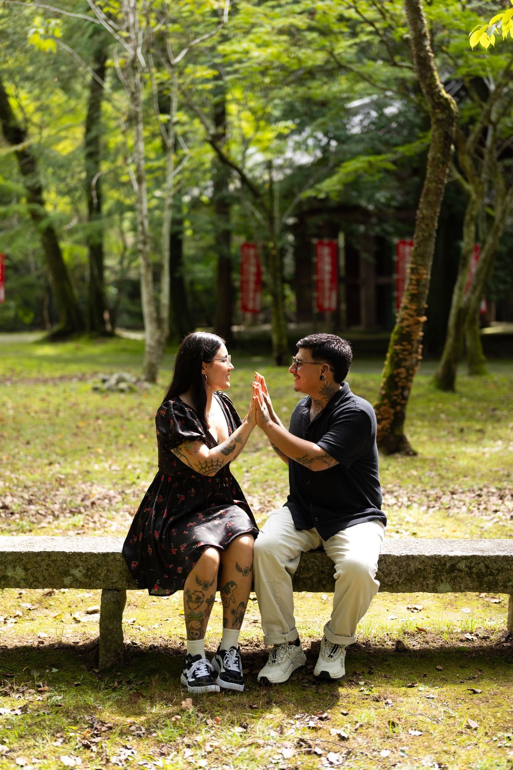 Tattooed couple sitting on a stone bench in a sunlit forest, pressing their palms together while smiling at each other.