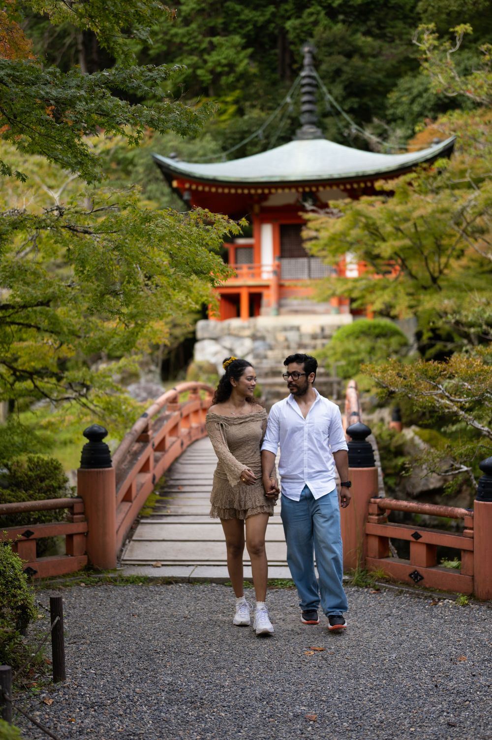 Couple holds hands while walking across a red wooden bridge toward a traditional Japanese pagoda surrounded by lush green trees.