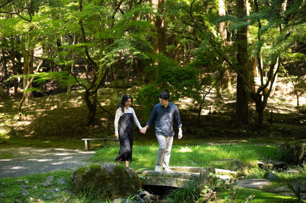 Couple holding hands while walking across a small stone bridge in a sunlit green forest park with tall trees and a bench in the background.
