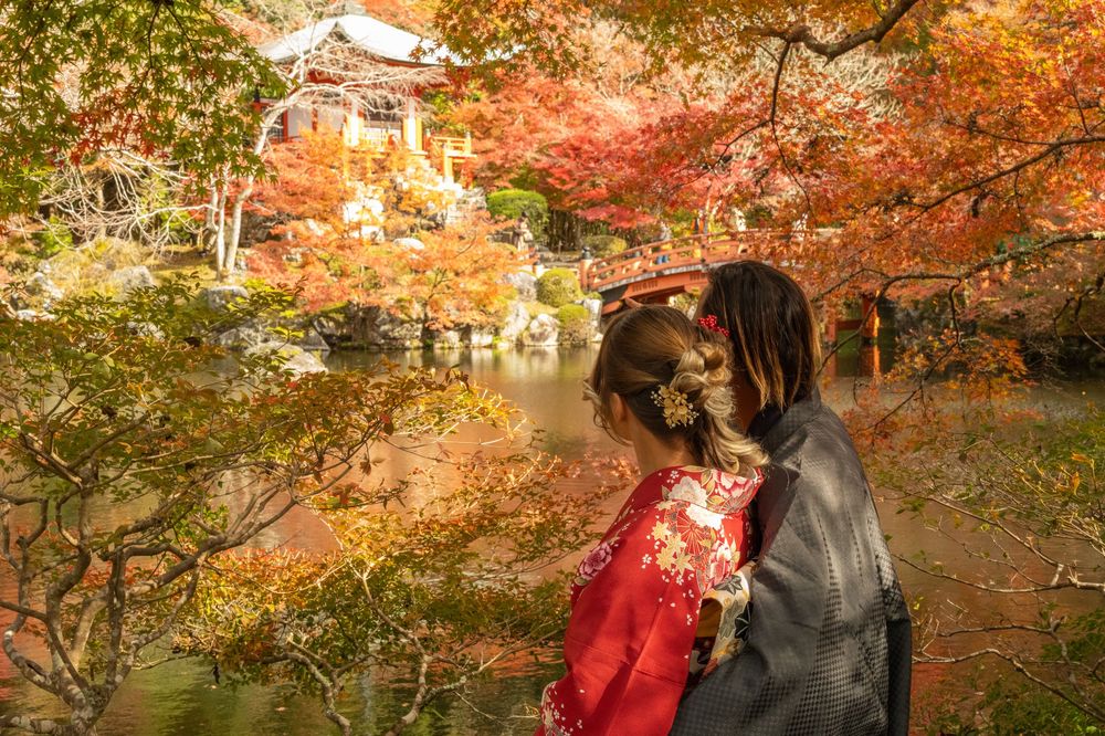 Two people in traditional Japanese kimono stand beside a pond surrounded by vivid red autumn maple leaves near a temple.