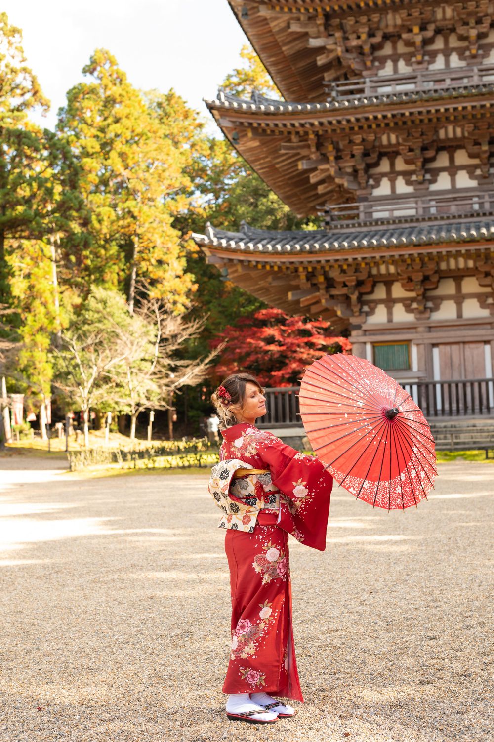 Woman in a red floral kimono holding a red oiled-paper parasol in front of a traditional Japanese pagoda surrounded by autumn trees.