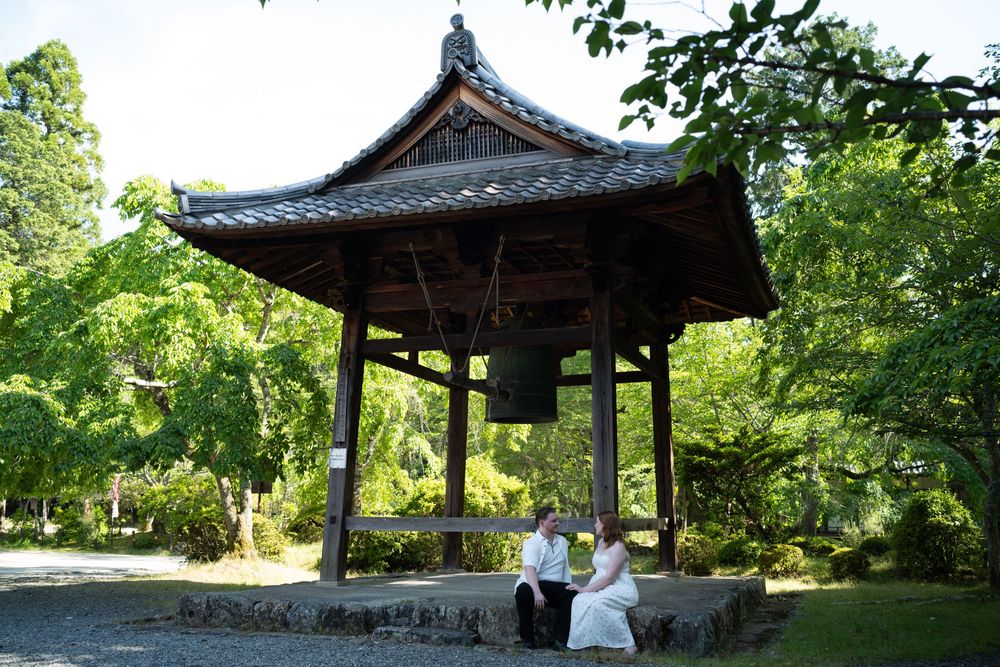 Couple in wedding attire sitting on the stone base of a traditional Japanese bell tower surrounded by lush green trees.