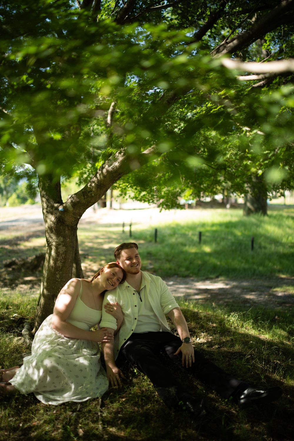 A couple sits close together beneath a large shady tree, the woman resting her head on the man's shoulder in a sunlit, grassy park.