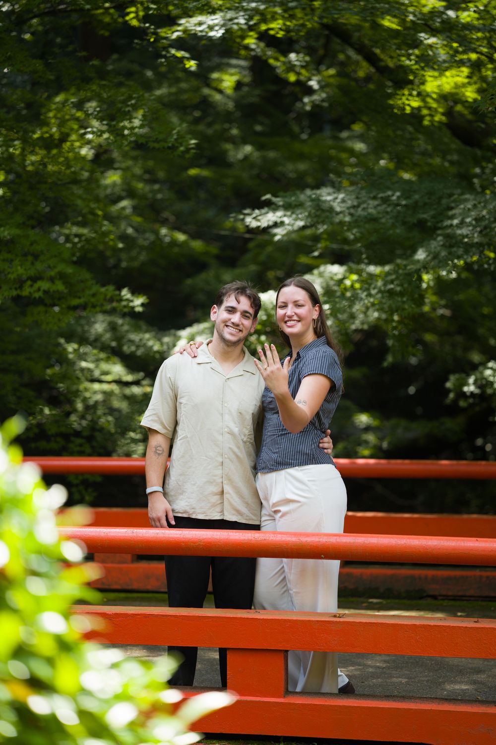 Smiling couple embracing on a red wooden bridge surrounded by lush green trees as the woman shows off her new engagement ring.
