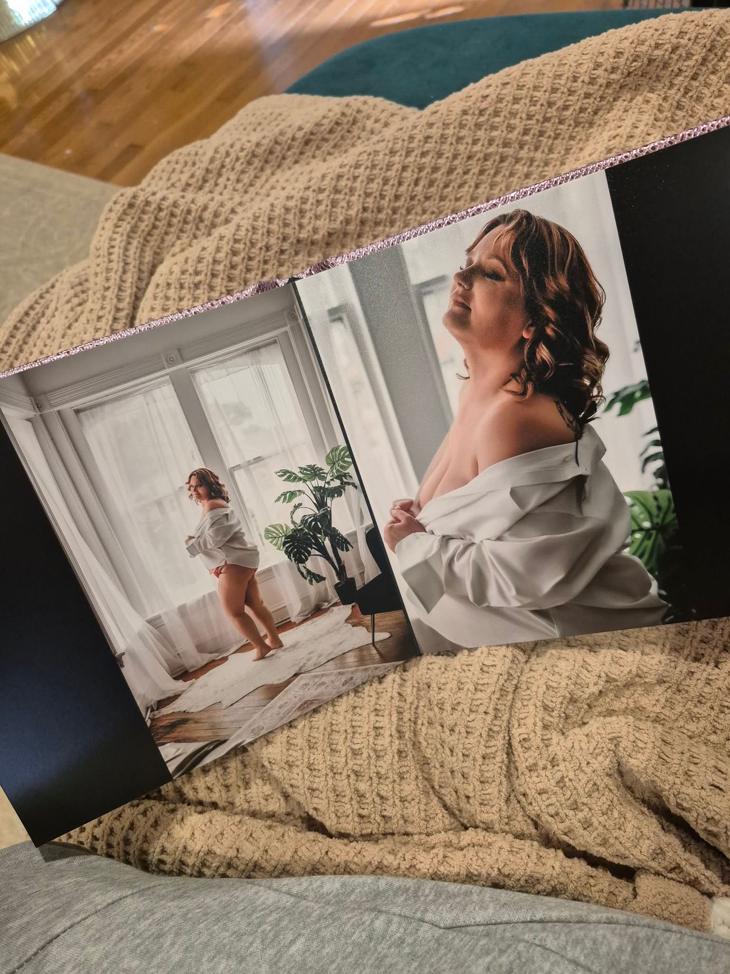 Open boudoir photo album on a knit blanket showing a woman in a white button-up shirt by a sunlit window and a full-length studio portrait.