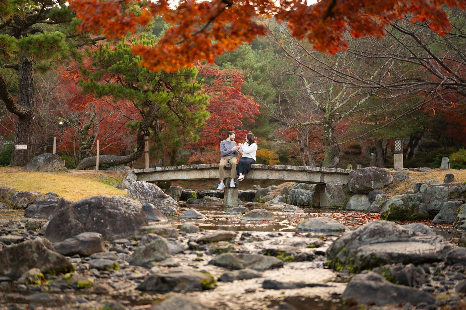 Two people sit on a stone bridge surrounded by vibrant autumn foliage in a serene Japanese garden.