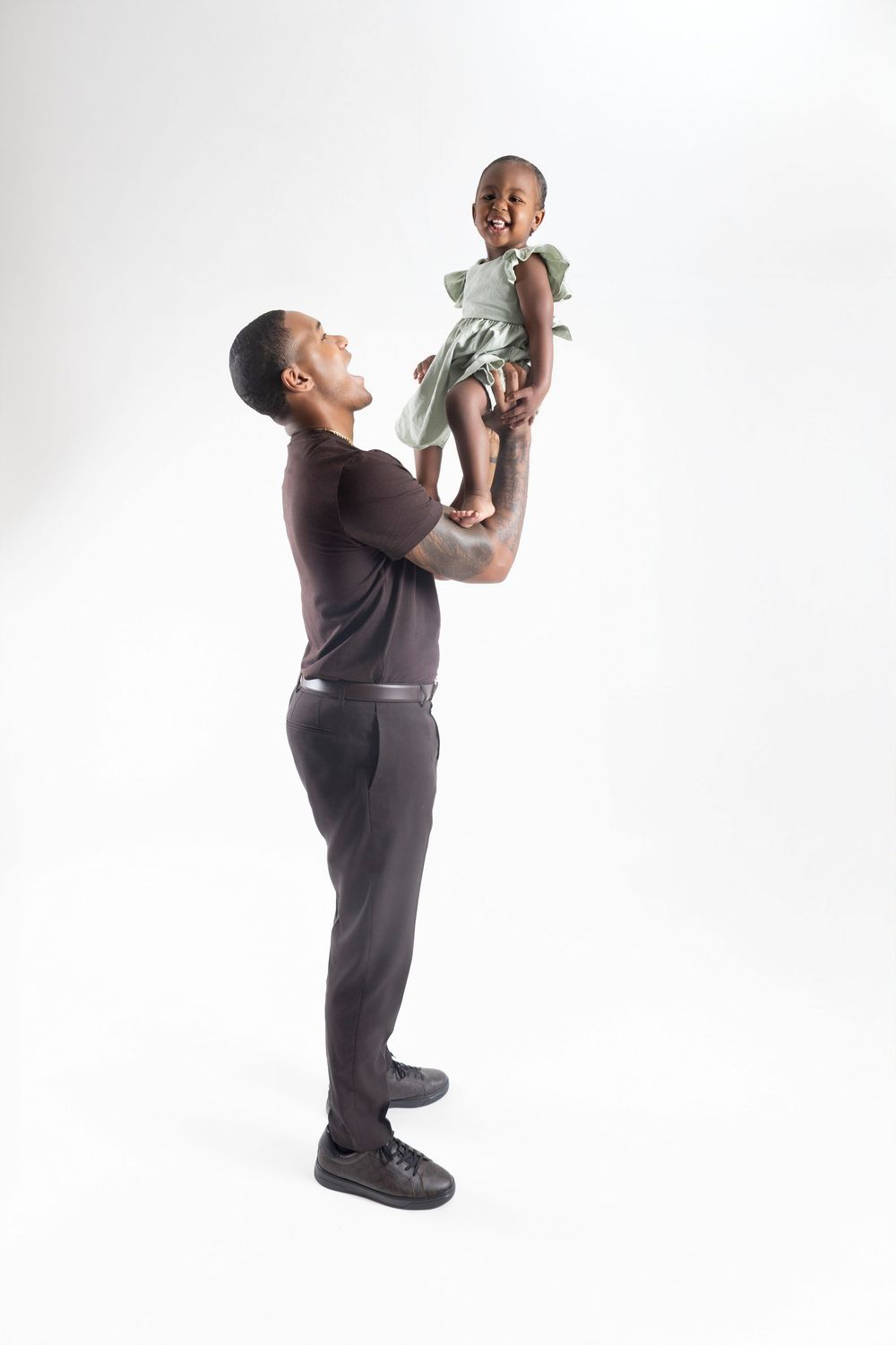 A father joyfully lifts his smiling baby girl in a green dress above his head against a white background.