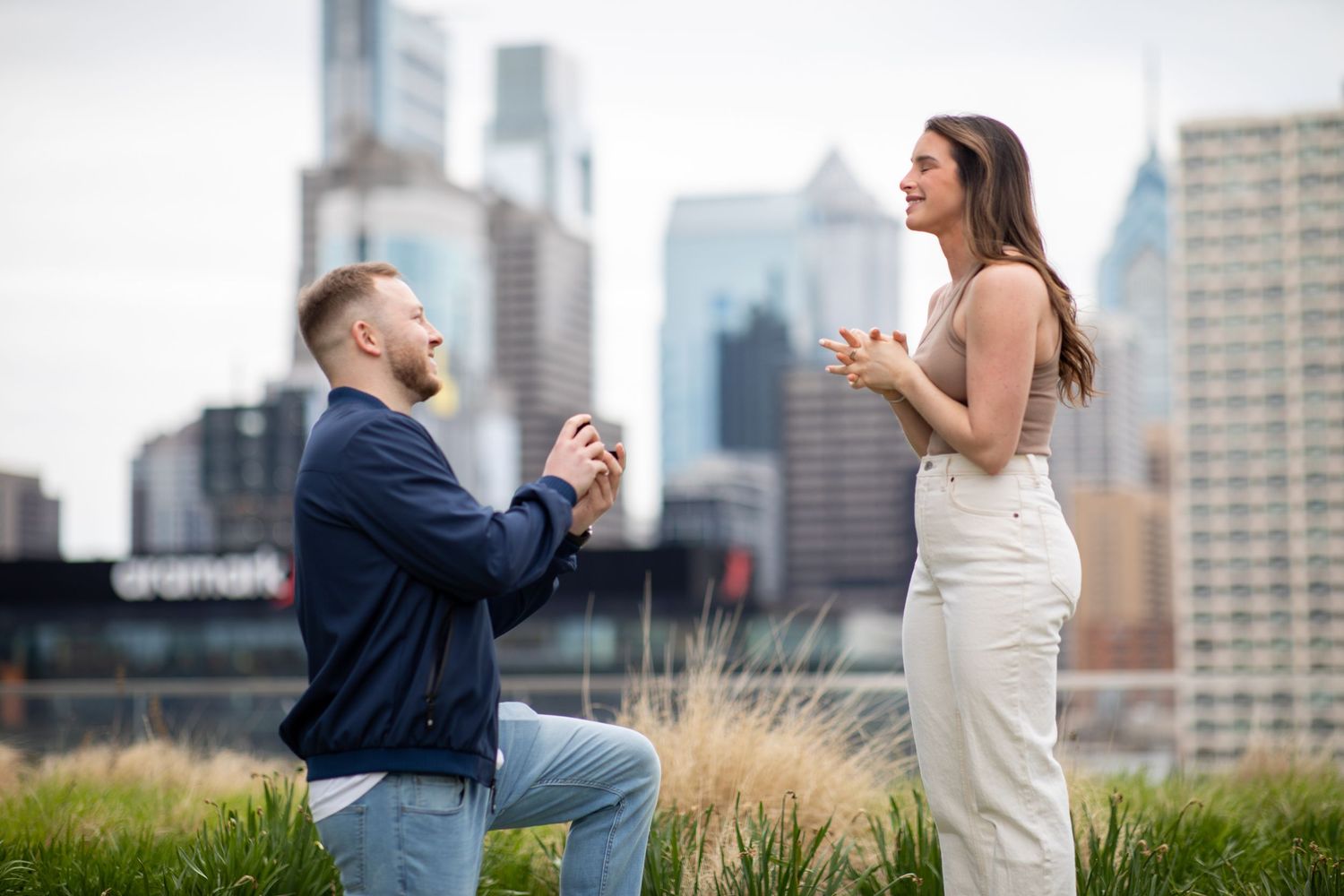 Man proposing to woman on one knee in urban park with Philadelphia city skyline in the background.