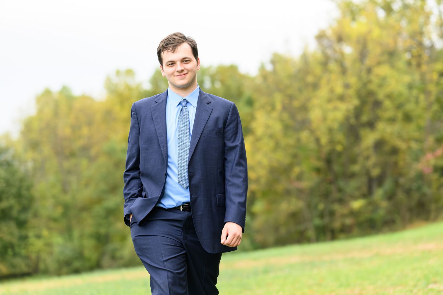 Young professional man in navy suit and light blue tie smiling outdoors in a green park setting.