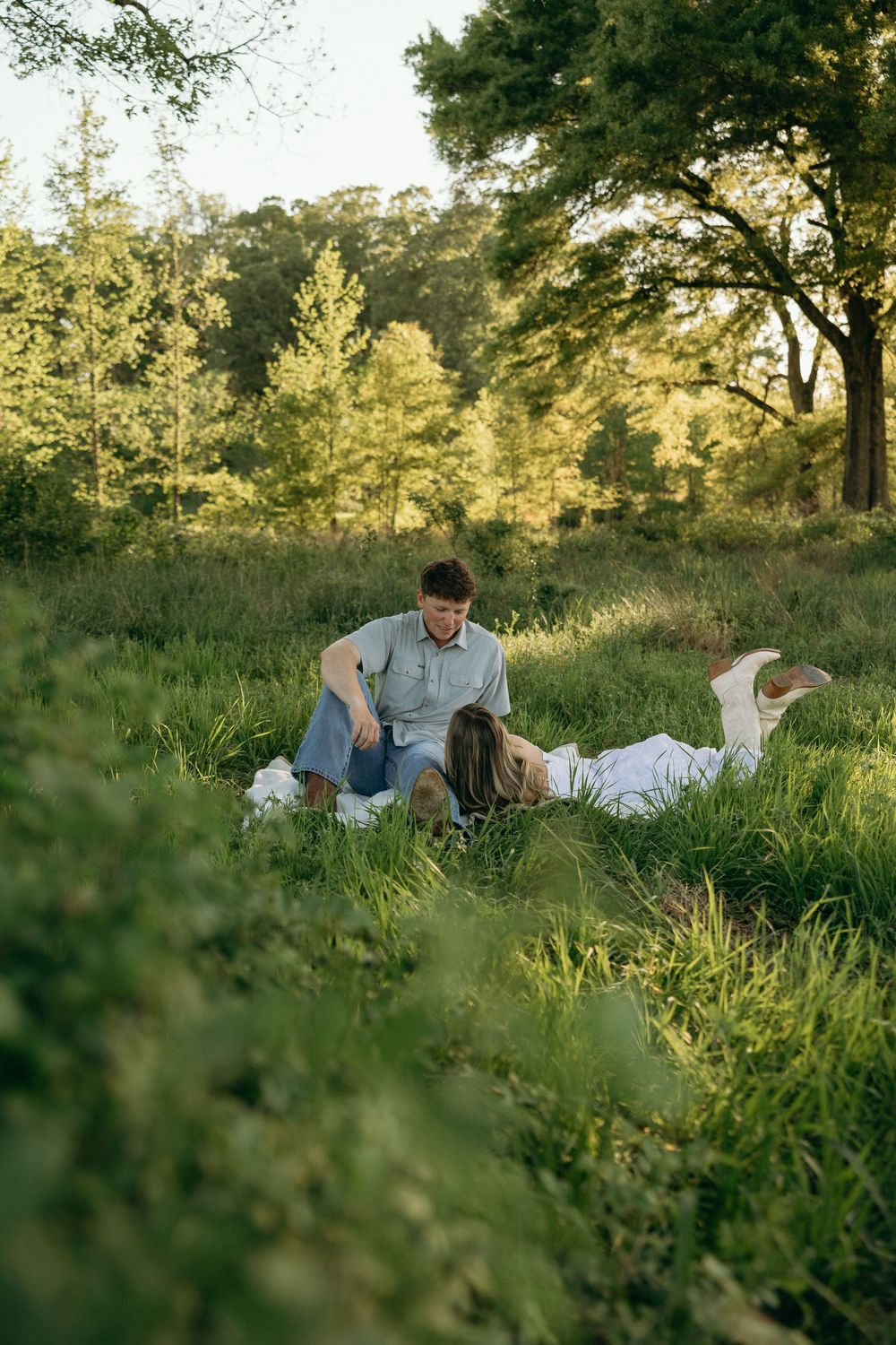 Couple relaxing in green meadow at golden hour, man sitting beside woman lying in tall grass near trees during outdoor engagement photos.