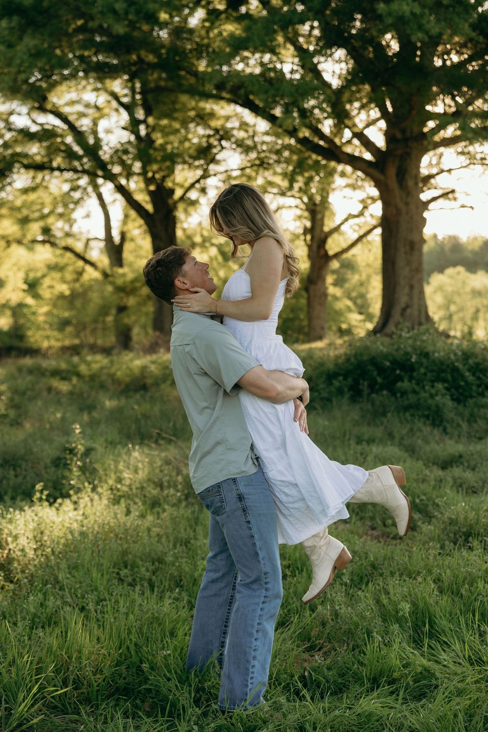 A man lifts a woman in a white dress for a photo during their engagement session on the farm near Atlanta.