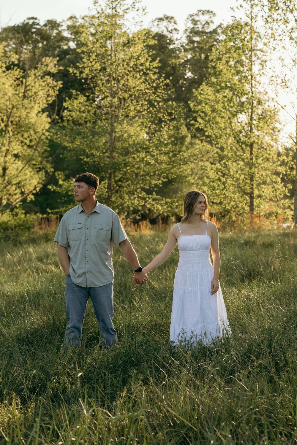 A couple holds hands in a sunlit green meadow, looking opposite directions during engagement session.