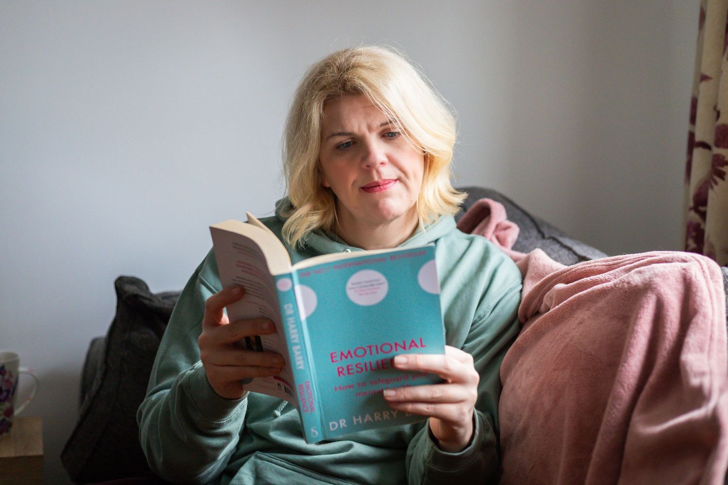 Woman in green sweater reading Emotional Design book while relaxing in a cozy chair with pink blanket.