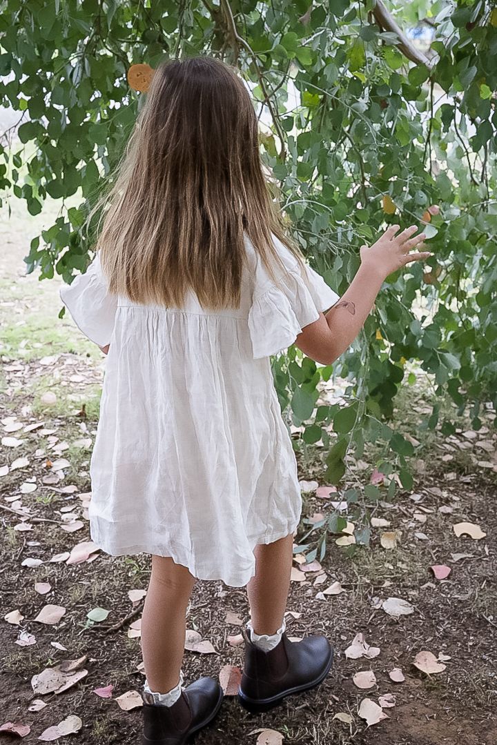 Young girl in white ruffled dress and brown boots reaching up to touch green tree leaves outdoors.
