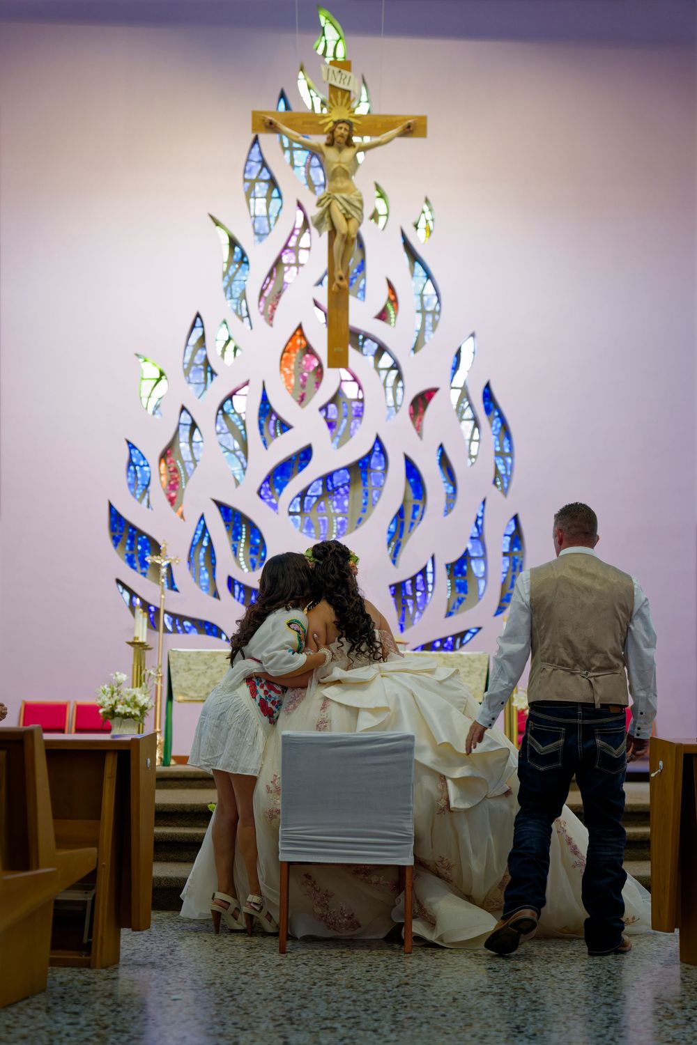 Young woman in white quinceañera gown kneels at church altar beneath ornate stained glass cross during religious ceremony.